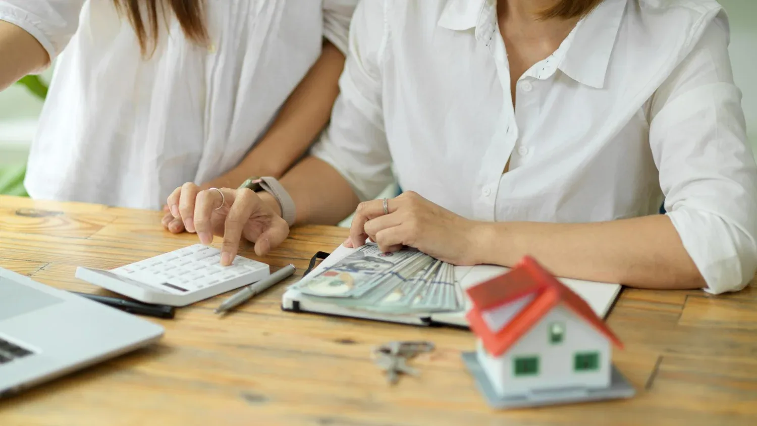 Duas mulheres sentadas à mesa usam calculadora para calcular valor de aluguel de imóvel residencial. Na mesa tem uma miniatura de casa, chaves, uma caderneta e dinheiro.